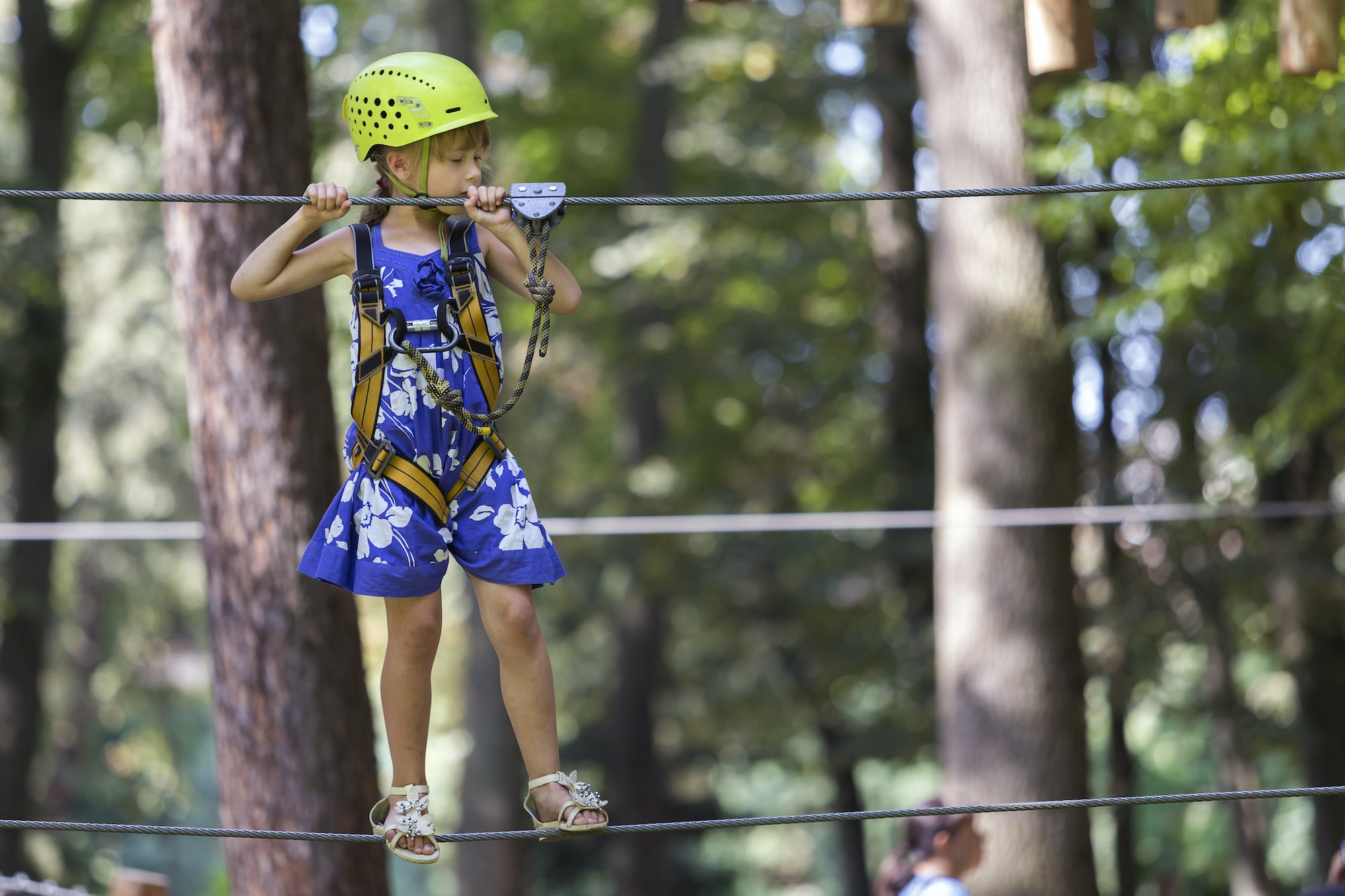 Young pretty blond child girl in safety harness and helmet on rope way on bright bokeh background.